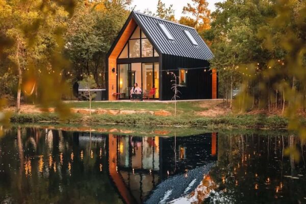 Mala moderna kuća s crnim krovom i velikim prednjim prozorom, uz jezero, okružena prirodom i svjetlucavim lampicama